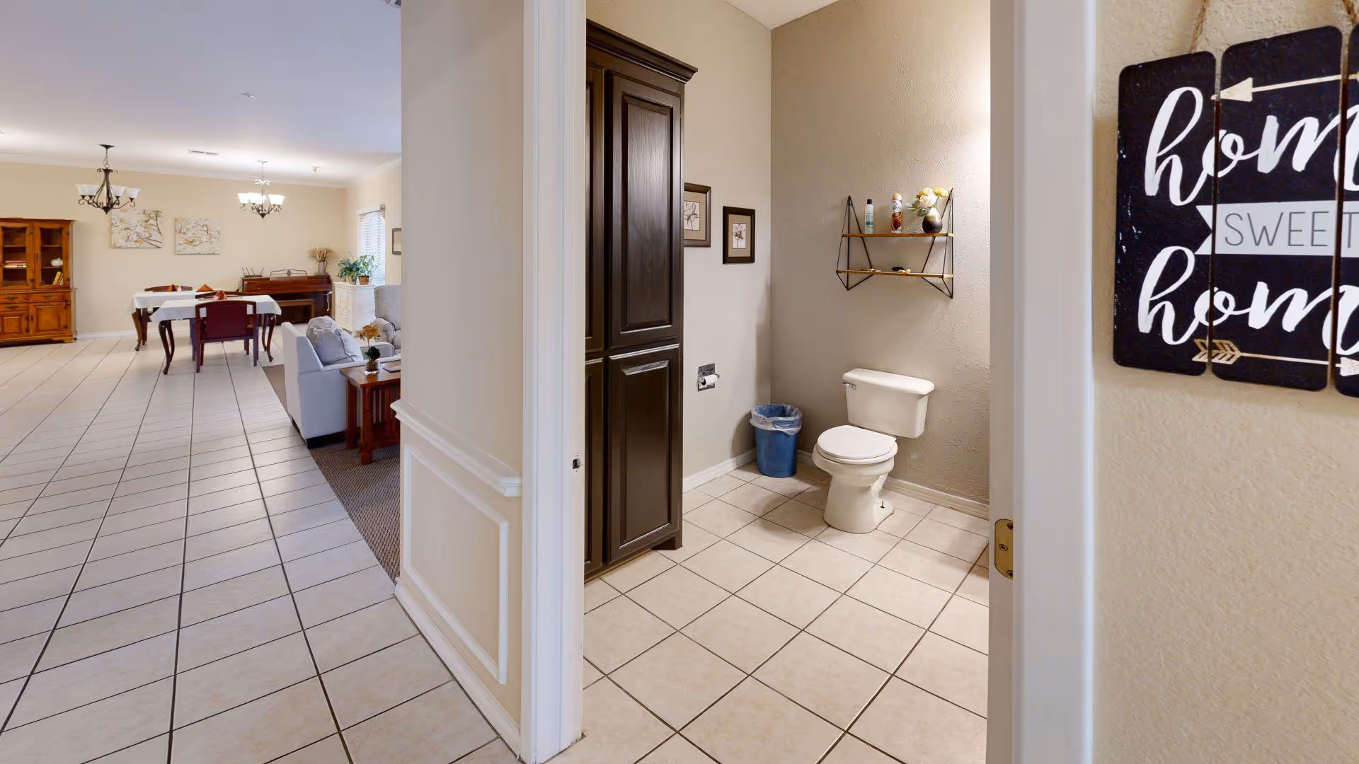 View of a tiled hallway leading to a dining area with a wooden dining table and chairs, and a living area with a sofa and piano. To the right, there is a small bathroom with a toilet, a blue trash bin, a dark wooden cabinet, and a wall shelf with decorative items. A sign on the wall near the bathroom door reads 'home sweet home'.