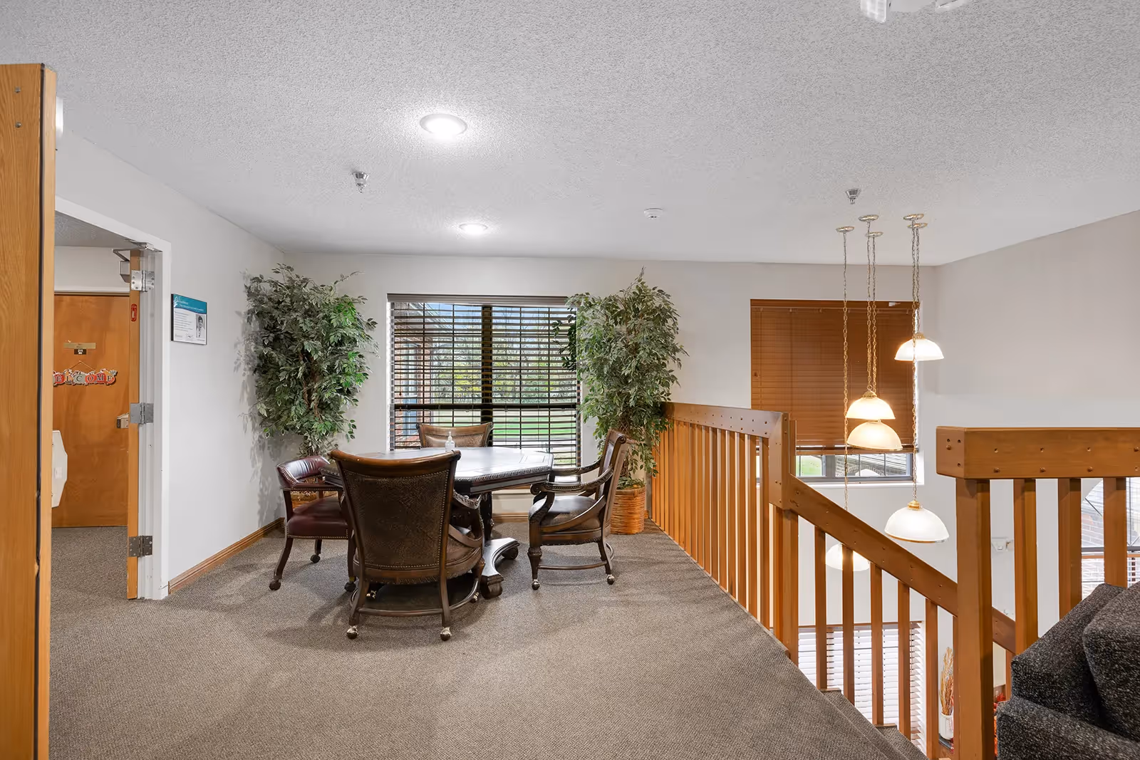Interior view of a senior living facility showing a small seating area with a round table and four chairs near a window with blinds. Two large potted plants are placed on either side of the window. The area is carpeted and has wooden railings overlooking a lower level with hanging pendant lights. A door is open to the left, revealing another room.