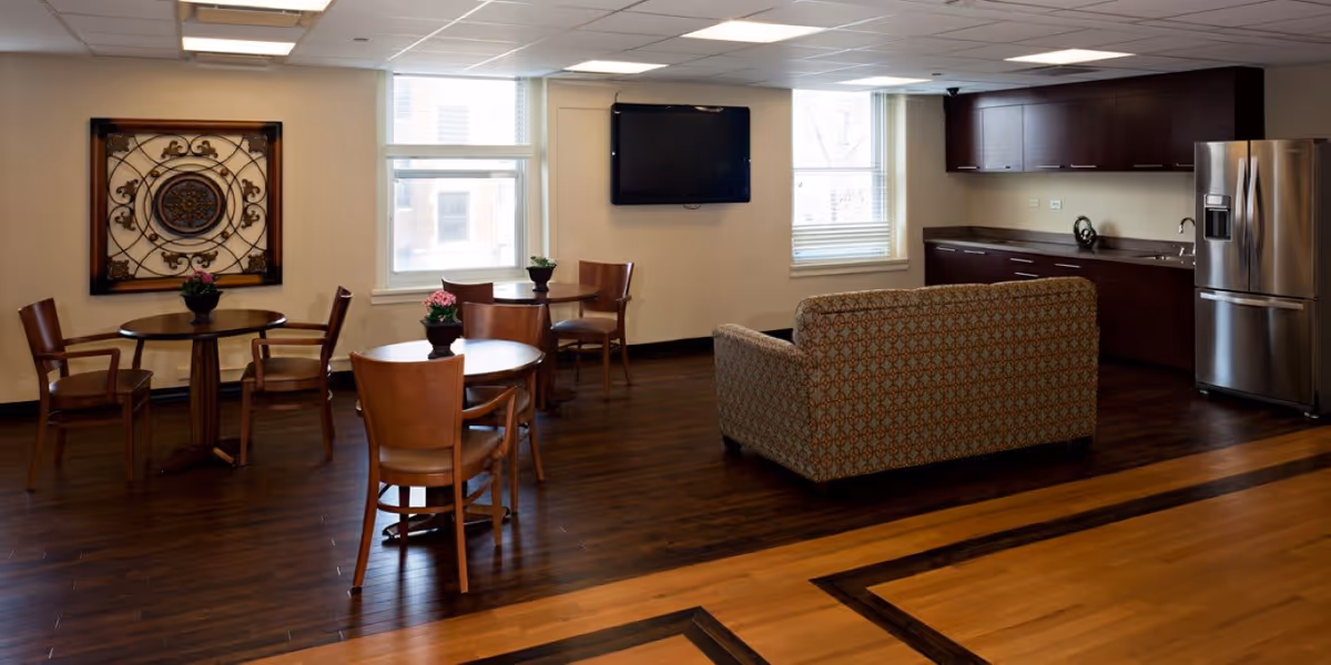 Communal lounge with round wooden tables and chairs, a patterned sofa facing a wall-mounted TV, and a kitchenette with a stainless steel refrigerator.