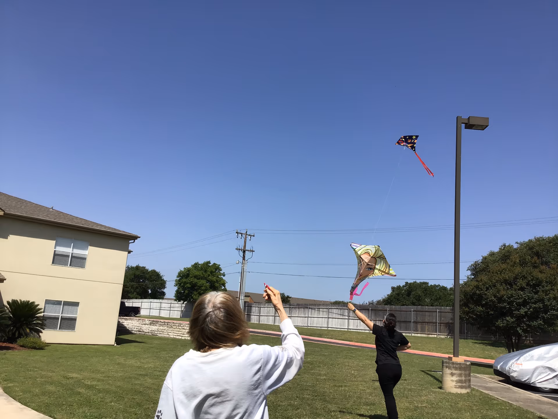 Two people flying kites in a grassy outdoor area near a senior living facility building under a clear blue sky. One person is wearing a white shirt and the other is in black. There are trees, a lamp post, and a covered vehicle in the background.