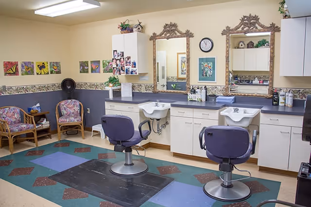 Interior view of a salon area in a senior living facility with two purple salon chairs in front of two sinks and mirrors. The room has floral patterned chairs, colorful artwork on the walls, and cabinets for storage.