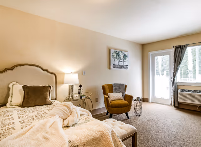 A cozy bedroom in an assisted living facility featuring a bed with a beige headboard, white and brown pillows, and a cream-colored blanket. Next to the bed is a small round glass side table with a lamp and a decorative item. There is a mustard yellow armchair with a pillow and a throw blanket, a small wire basket, a wall painting of flowers, and a door with a window leading outside. The room has beige walls and carpeted floor.