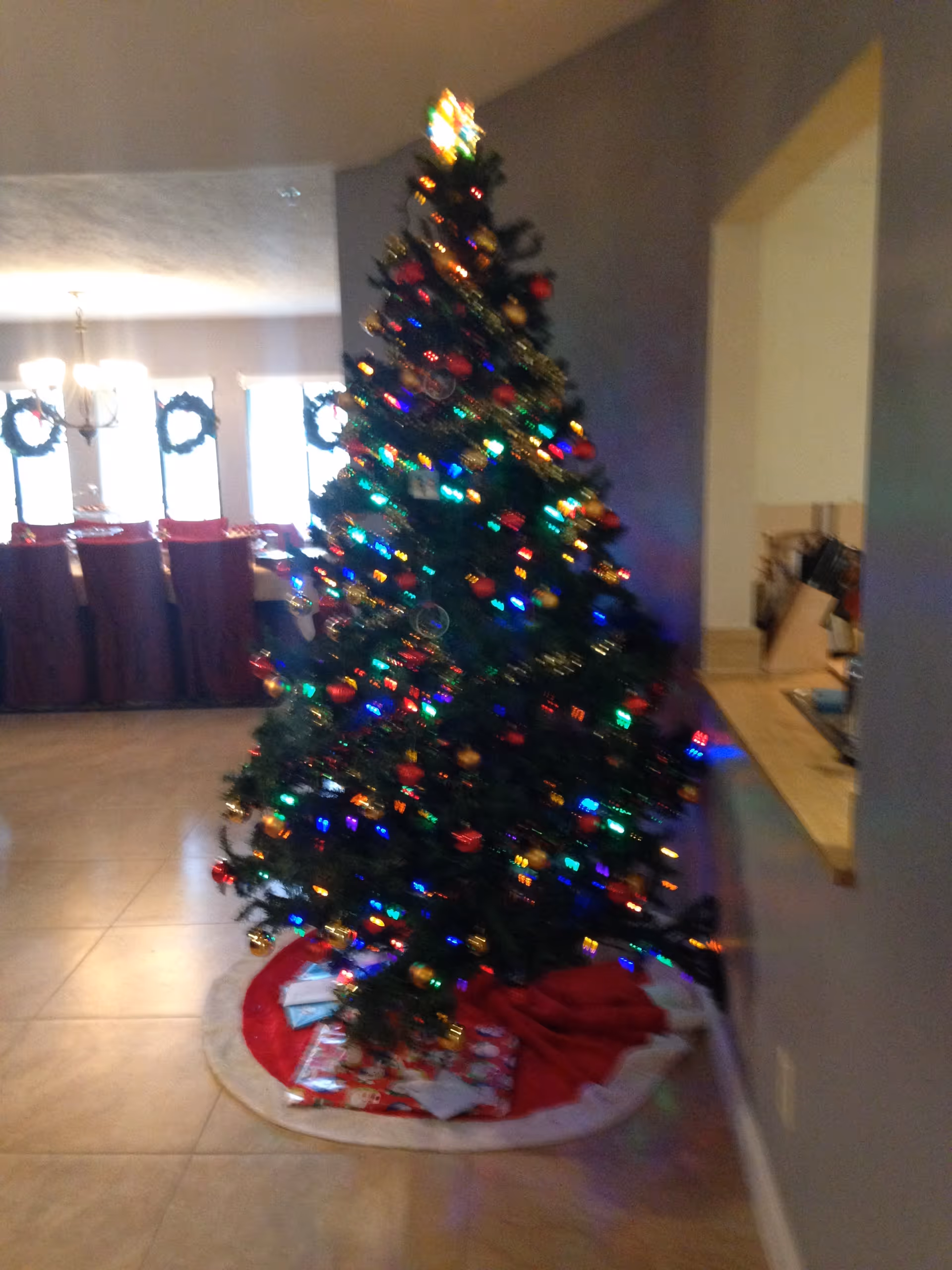 A decorated, brightly lit Christmas tree with presents beneath stands in an open living area.