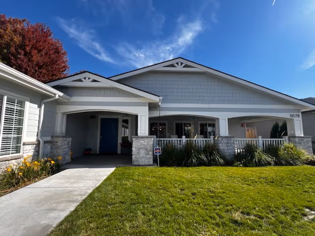 Single-story building front with a covered entrance, stone columns, white porch railing, green lawn, and blue sky above.
