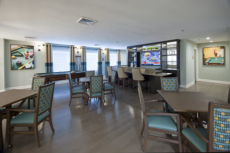 A spacious common area in Allegro Senior Living featuring multiple tables and chairs with patterned upholstery, a bar counter with stools, a wall-mounted TV, and two paintings depicting billiards scenes. Large windows with curtains allow natural light to brighten the room.