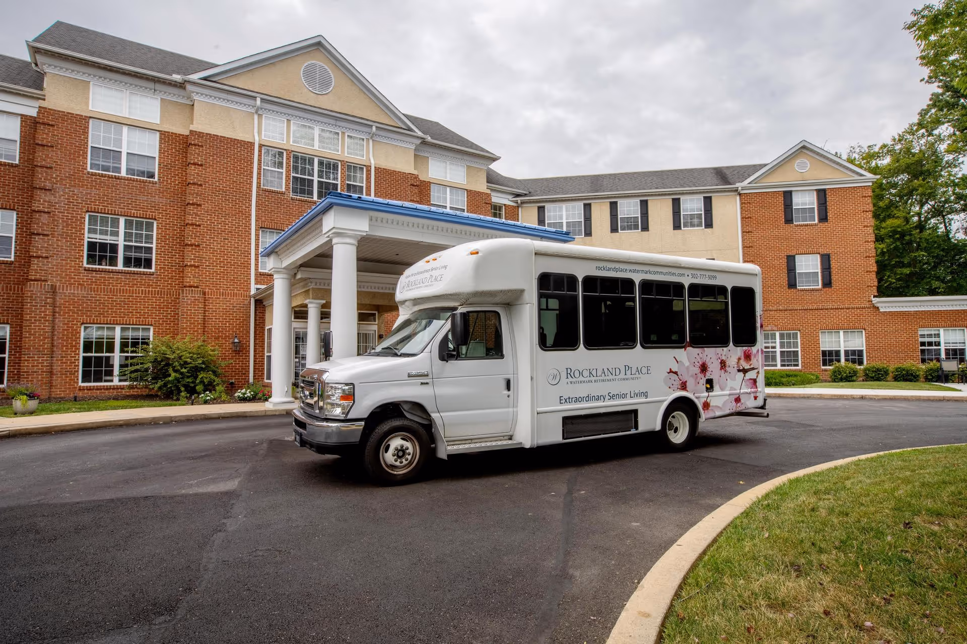 A white shuttle bus with floral designs and the text 'Rockland Place Extraordinary Senior Living' parked in front of a large brick and beige senior living facility building with multiple windows and a covered entrance with white columns.
