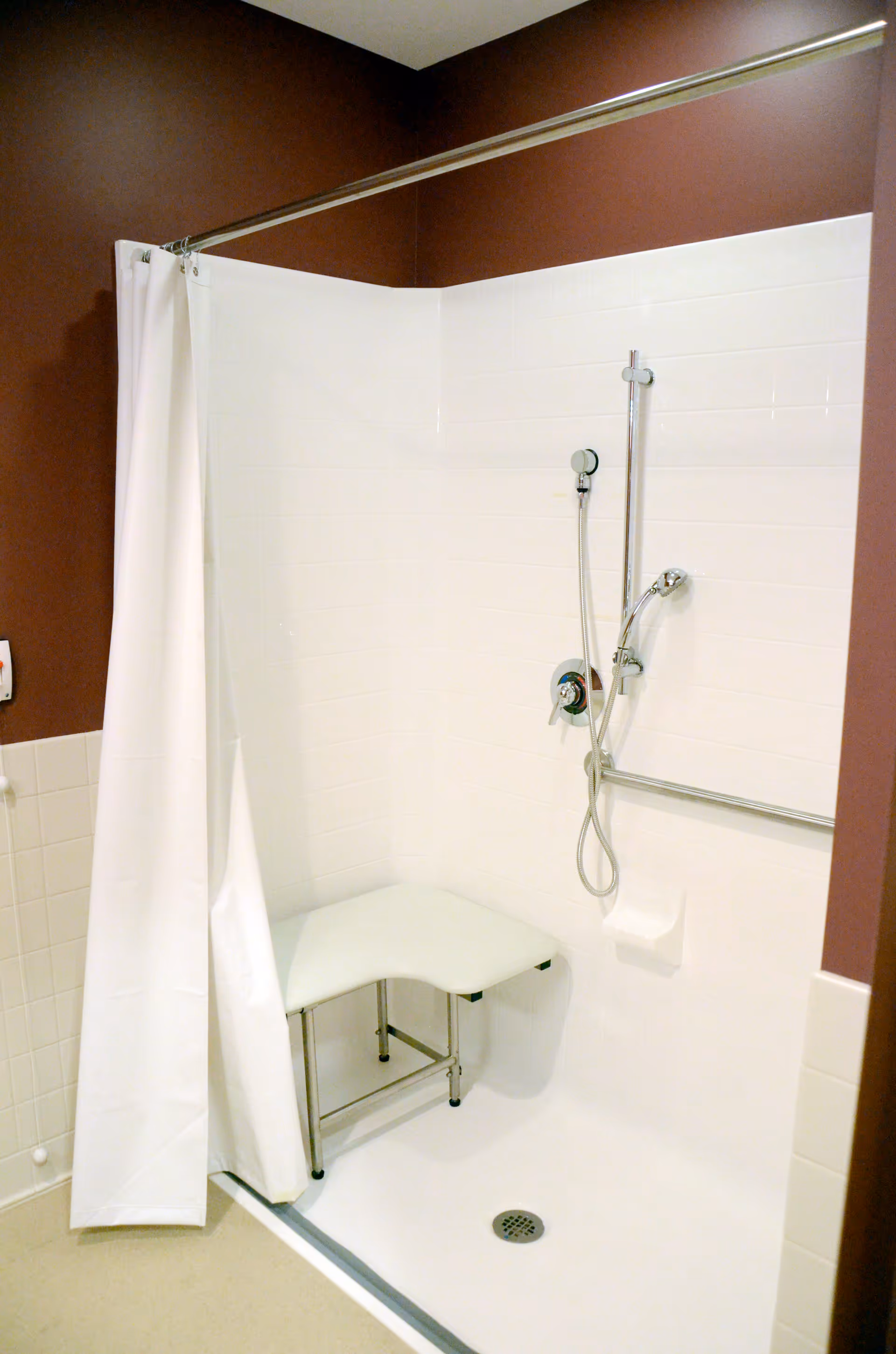 Accessible tiled shower stall with a white curtain, corner bench, handheld showerhead, and grab bar.