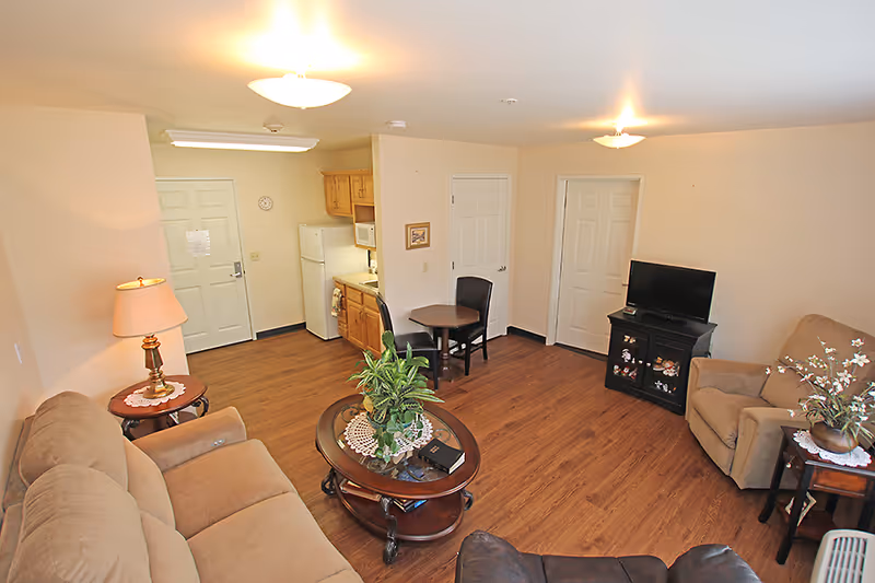 Bright open living room with sofa, armchairs, coffee table, TV stand and a small kitchenette in the background.