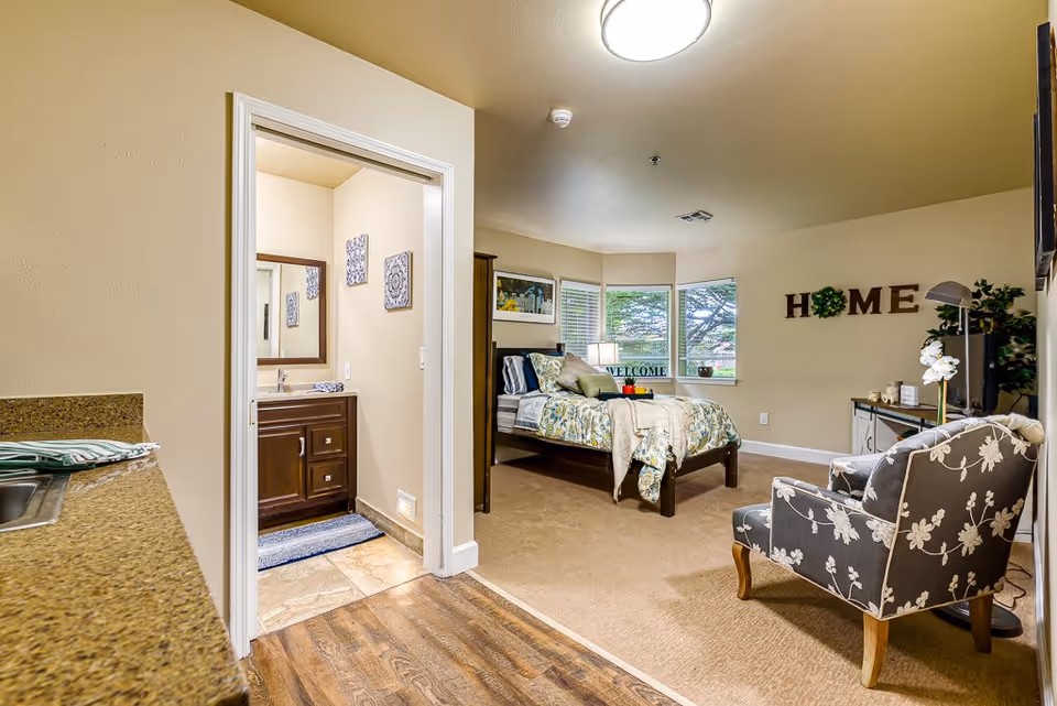 Interior view of a senior living facility room at The Cottages of Snohomish showing a cozy bedroom with a bed, decorative pillows, a chair with floral upholstery, a TV on a stand, and a wall decoration spelling 'HOME'. To the left, there is a small bathroom with a sink and mirror visible through an open door, and a kitchenette countertop with a sink is partially visible in the foreground.