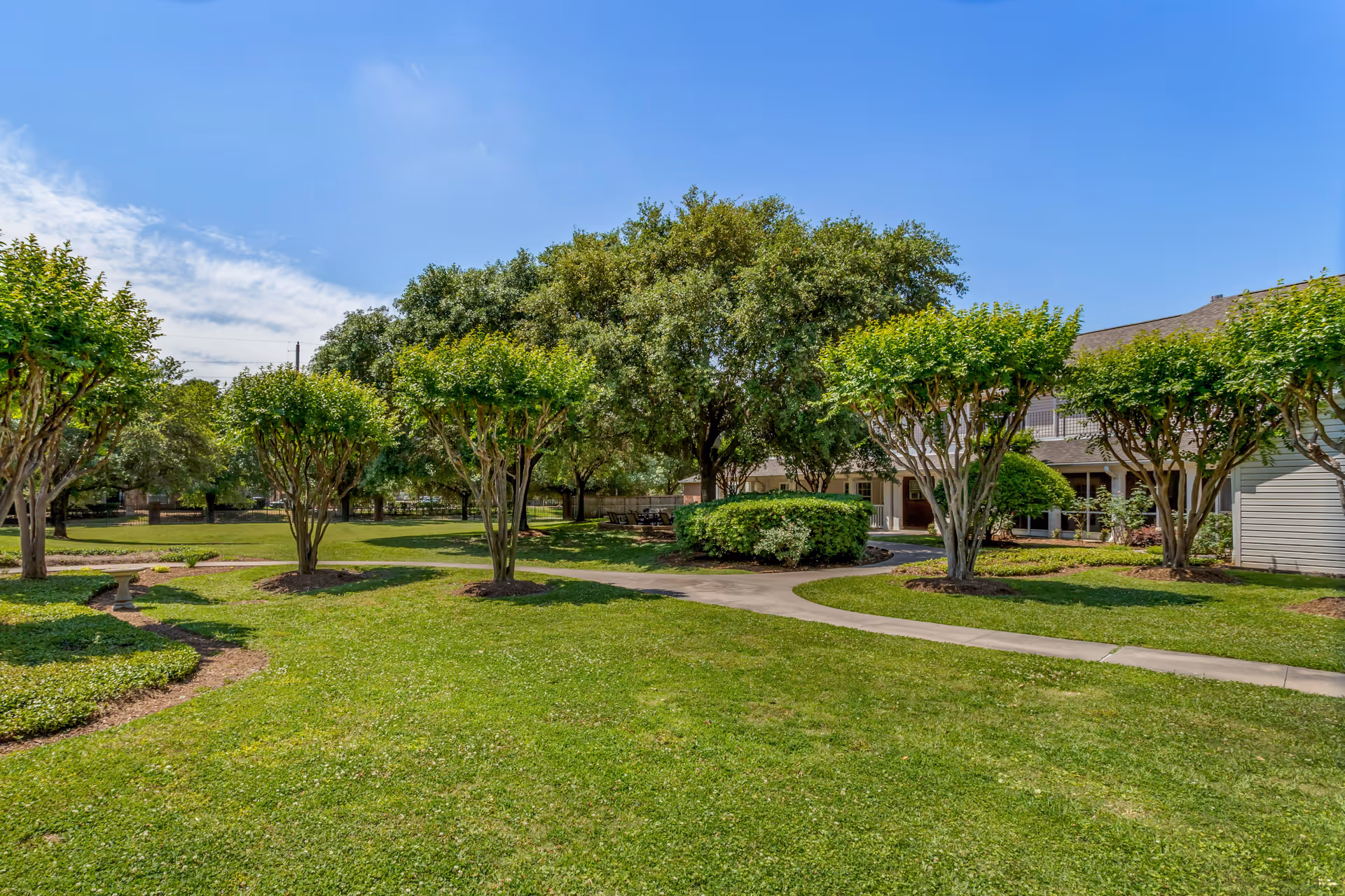 A well-maintained outdoor garden area at Brookdale Shadowlake with neatly trimmed trees, green grass, a paved walkway, and a building partially visible in the background under a clear blue sky.