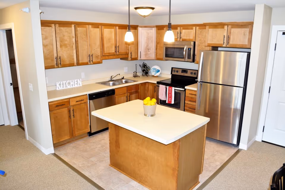 A modern kitchen with wooden cabinets, stainless steel appliances including a refrigerator, microwave, dishwasher, and stove. There is a kitchen island in the center with a bowl of lemons on top. Two pendant lights hang from the ceiling above the island. The floor is tiled and the walls are painted light beige.