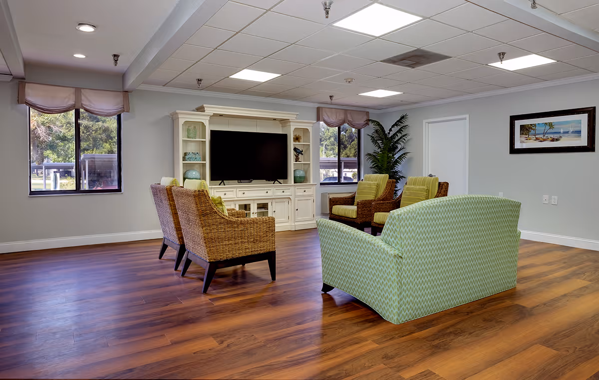 A spacious living room with wooden flooring, featuring a green patterned sofa, four wicker armchairs with green cushions arranged around a large flat-screen TV on a white entertainment center. The room has two windows with light pink valances, a large potted plant, and a framed beach-themed picture on the wall.