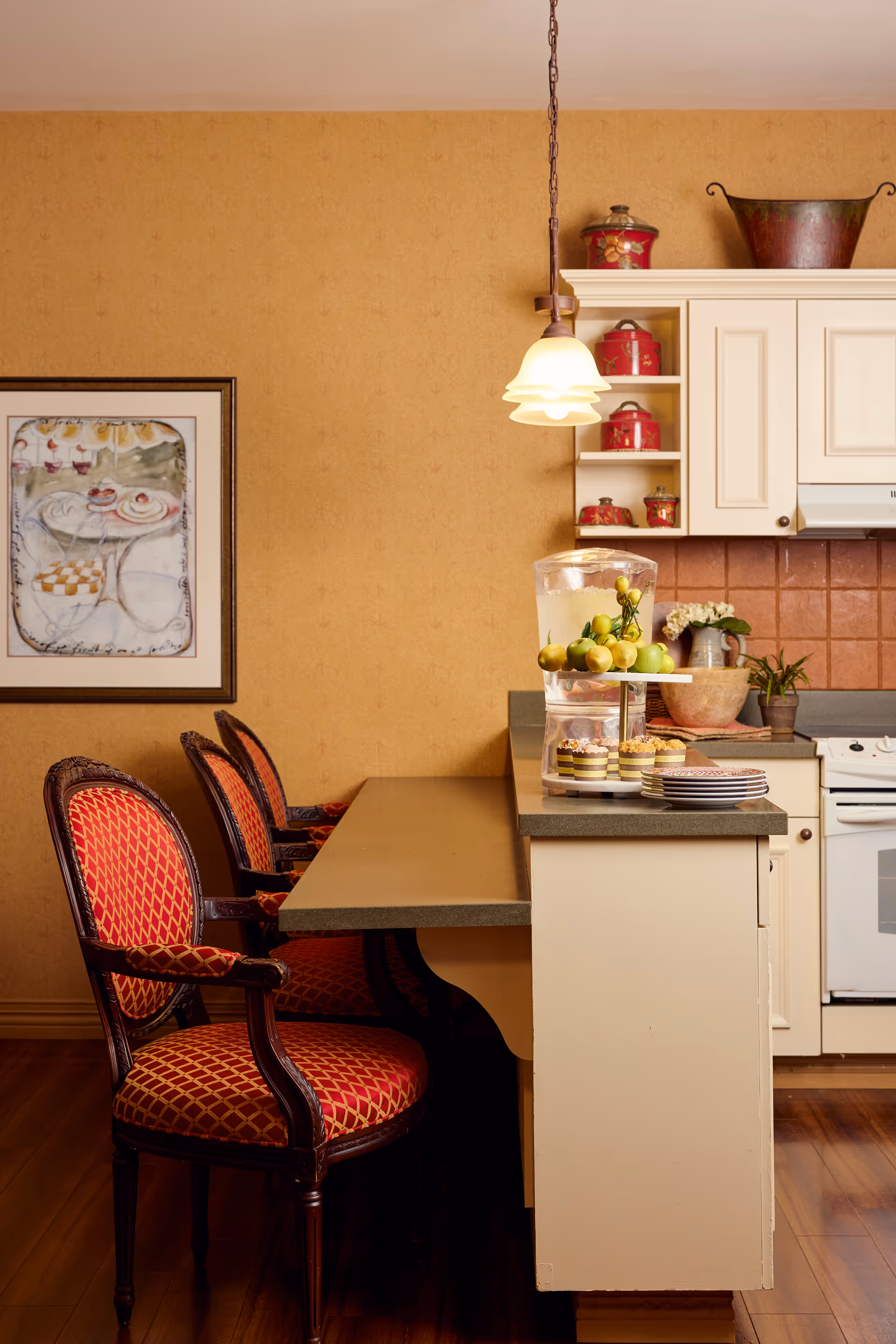 A cozy kitchen area with a countertop and three red cushioned chairs with wooden frames. On the countertop, there is a glass container with water and lemons, a tiered tray with cupcakes, and a stack of plates. The kitchen cabinets are cream-colored with red decorative containers on the shelves. A pendant light hangs above the counter, and a framed artwork is on the wall.
