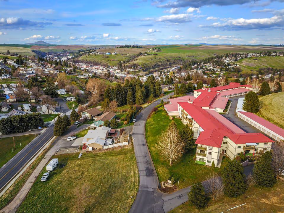 Aerial view of a residential area with a large building complex featuring red roofs, surrounded by green fields, trees, and a winding road. The landscape includes houses, roads, and rolling hills under a partly cloudy sky.