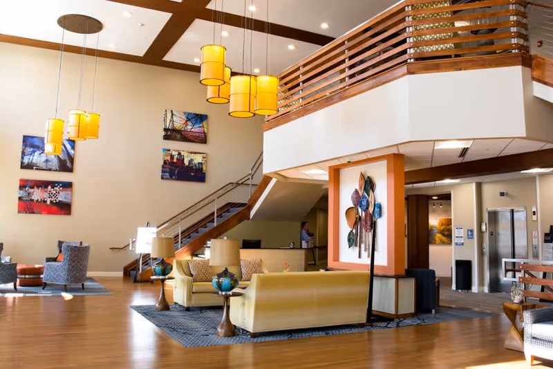 Spacious senior living facility common area with wooden flooring, modern yellow sofas, blue patterned armchairs, and hanging yellow pendant lights. A staircase with wooden railings leads to an upper level. Colorful abstract artwork is displayed on the walls, and there is a decorative wall piece with multicolored leaves near the staircase. An elevator is visible in the background.