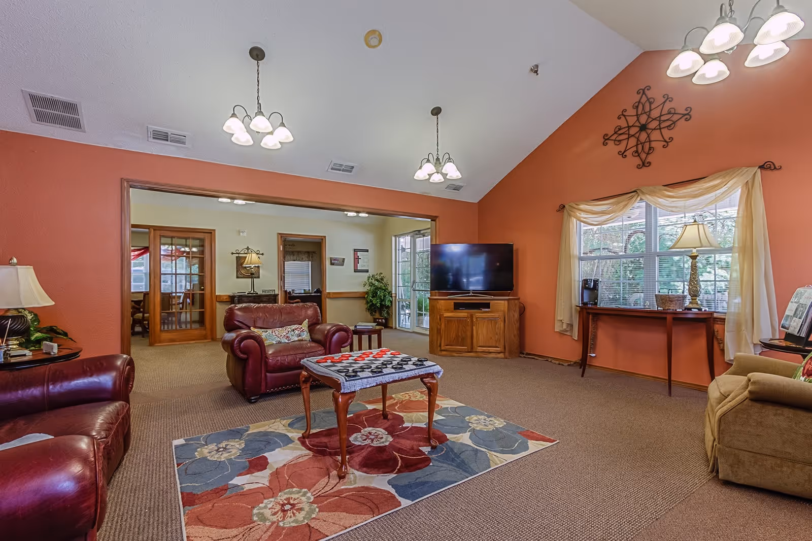 A cozy living room with red and beige walls, featuring two red leather armchairs, a beige armchair, a wooden coffee table with a checkers game on it, a colorful floral rug, a flat-screen TV on a wooden stand, and a window with sheer curtains and a lamp on a console table beneath it.
