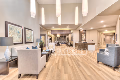 A spacious and well-lit senior living facility common area with light wood flooring, modern pendant lights hanging from a high ceiling, comfortable armchairs arranged around small tables, decorative framed artwork on the walls, and a long hallway leading to other rooms.