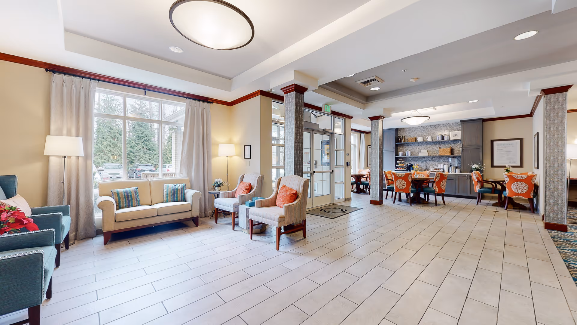 A bright and spacious common area in a senior living facility featuring a seating area with a beige sofa, two armchairs with orange cushions, and two teal armchairs near a large window with curtains. The room has tiled flooring, a ceiling light fixture, and a small dining area with orange patterned chairs and a cabinet with shelves holding various items. The entrance door is visible in the background.