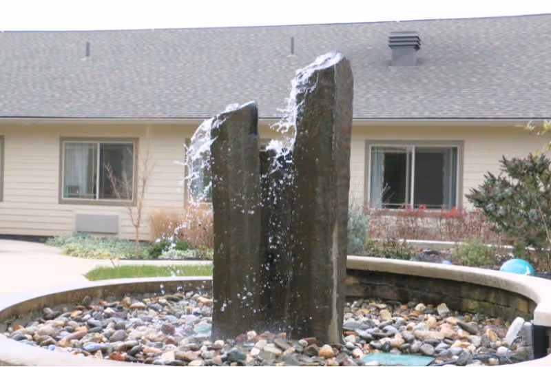 Outdoor water fountain with two vertical stone pillars spouting water, surrounded by a circular stone basin filled with small rocks. In the background, there is a beige building with windows and some landscaping including bushes and small trees.