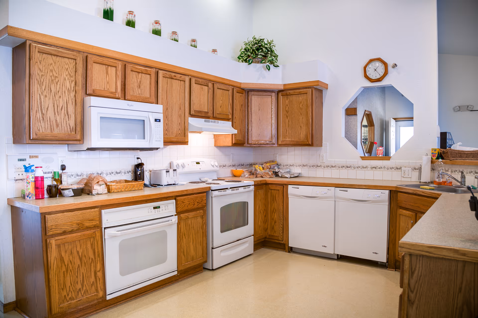 A bright kitchen with wooden cabinets, white appliances including a microwave, stove, oven, and two dishwashers. The countertop has various items such as a toaster, bread, and kitchen supplies. There is a small octagonal window on the wall and a clock above it. The kitchen has a clean and organized appearance.