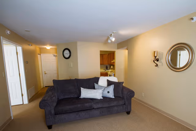 A cozy living room area with a dark gray sofa adorned with three decorative pillows. The room has beige walls and carpeted flooring. On the wall to the right, there is a round mirror and a wall-mounted candle holder. In the background, a kitchen area with wooden cabinets is partially visible. A clock is mounted on the wall near the kitchen entrance, and a hallway with white doors is seen on the left side.