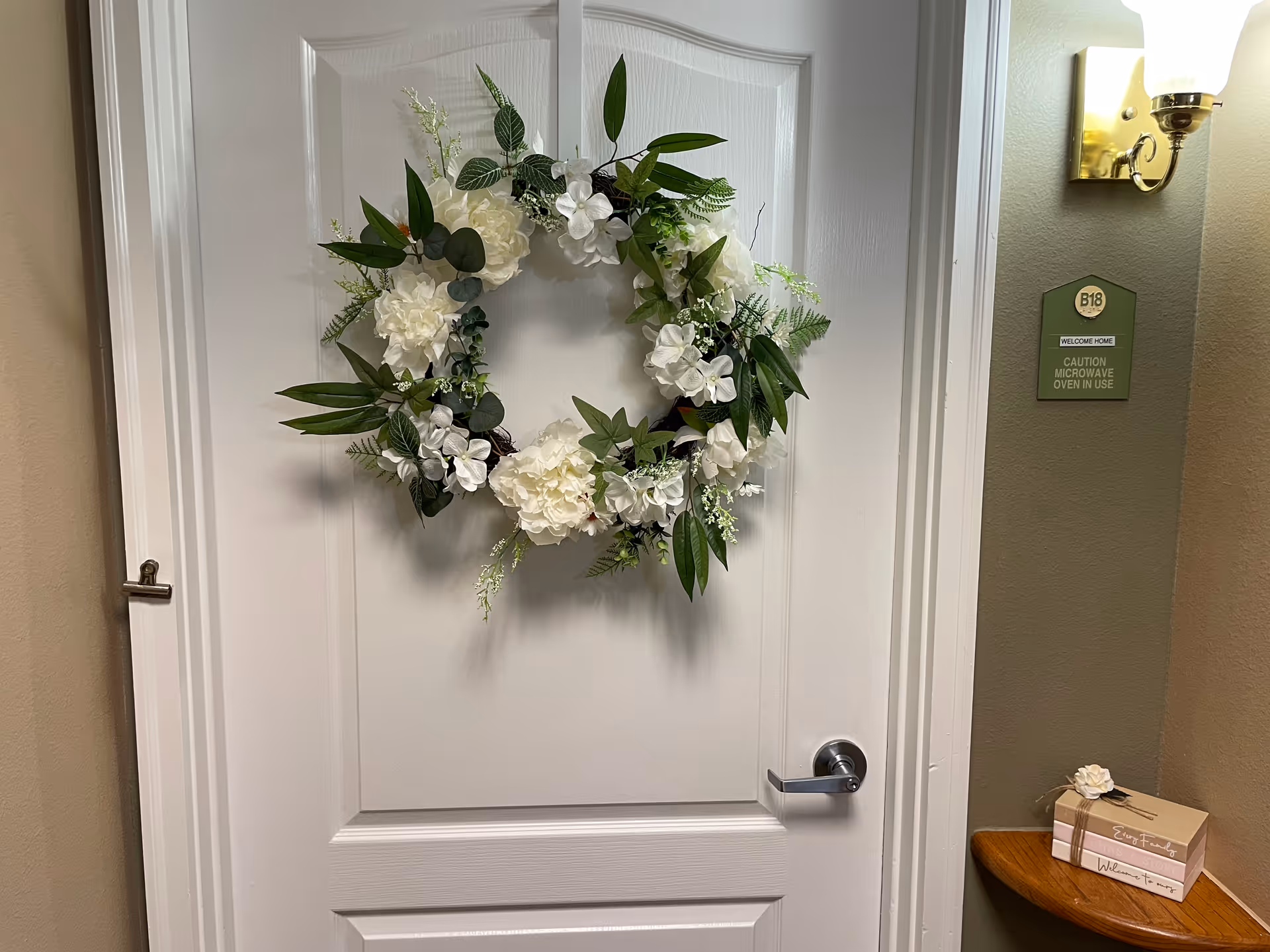 A white door decorated with a wreath made of white flowers and green leaves. To the right of the door is a green wall with a light fixture above and a sign that reads 'B18 WELCOME HOME CAUTION MICROWAVE OVEN IN USE'. Below the sign is a small wooden shelf holding a decorative box with a white flower on top.