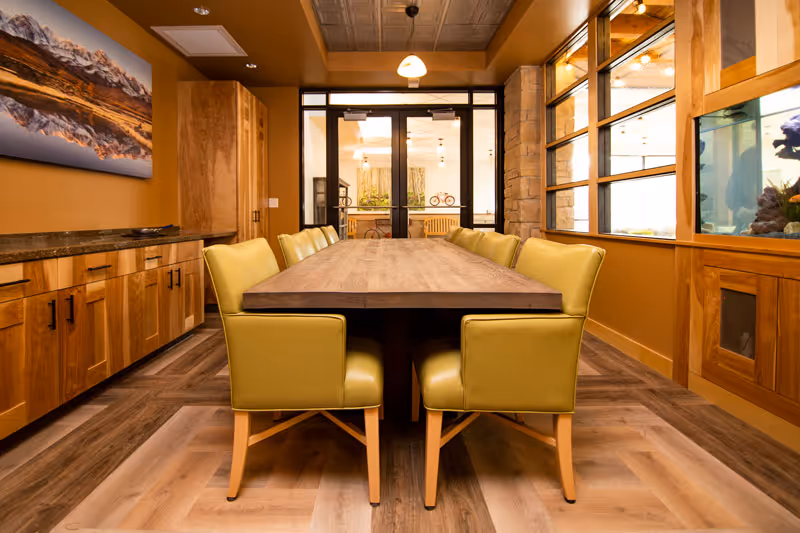 A dining room with a long wooden table surrounded by eight yellow cushioned chairs. The room features wooden cabinetry along one wall, a large window with wooden framing on the right, and a painting of a mountain landscape on the left wall. The floor has a wood pattern, and there is a glass door at the far end leading to another room.
