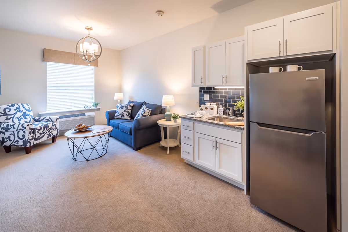 Bright studio-style living area with a small kitchenette, blue sofa, patterned armchair, coffee table, and stainless steel refrigerator.