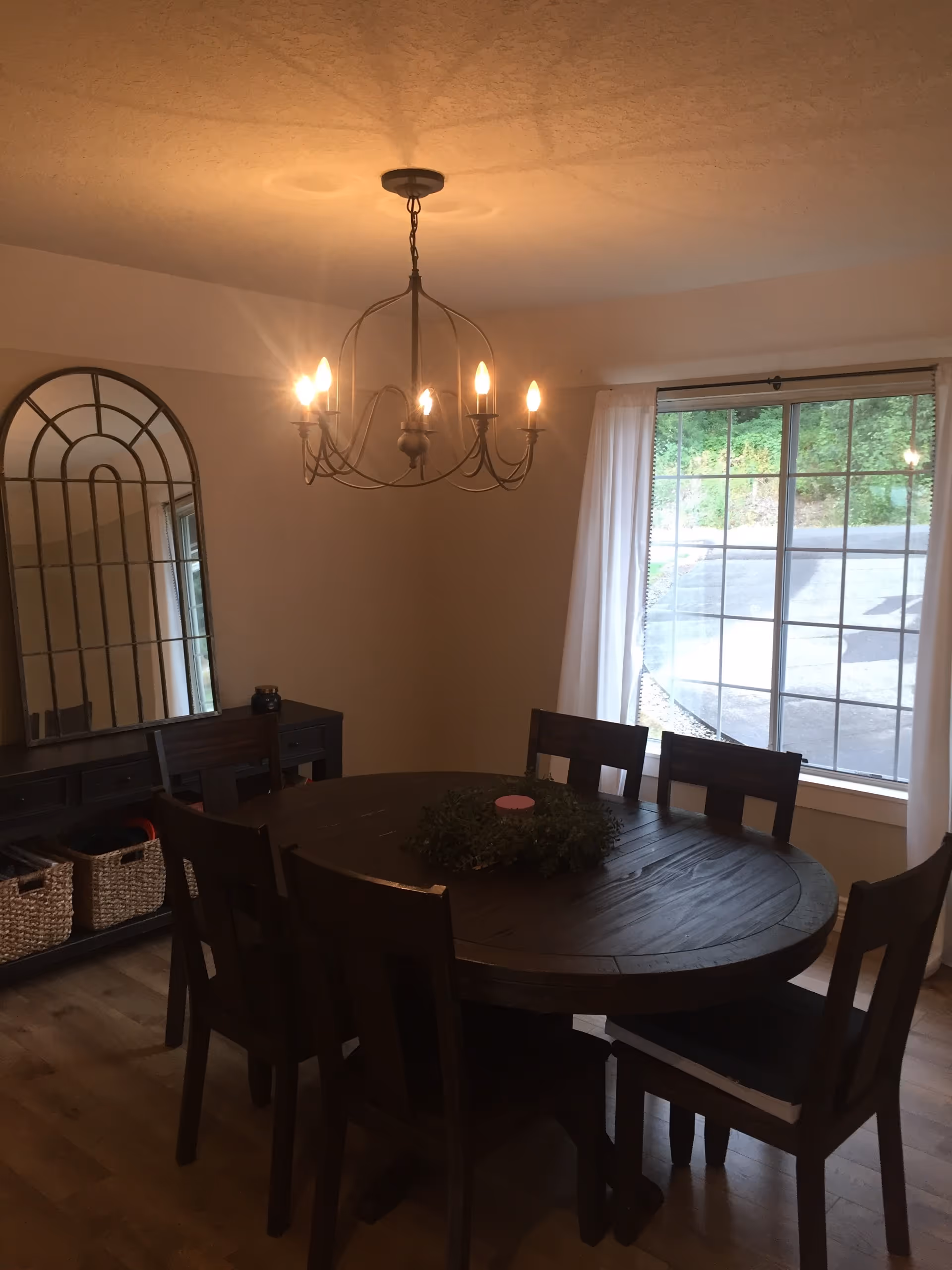A dining room with a round wooden table surrounded by six wooden chairs. A decorative wreath with a pink candle is placed in the center of the table. A chandelier with six lit bulbs hangs from the ceiling above the table. There is a large window with white curtains letting in natural light, and a dark wooden sideboard with woven baskets underneath and a large arched mirror on the wall behind it.