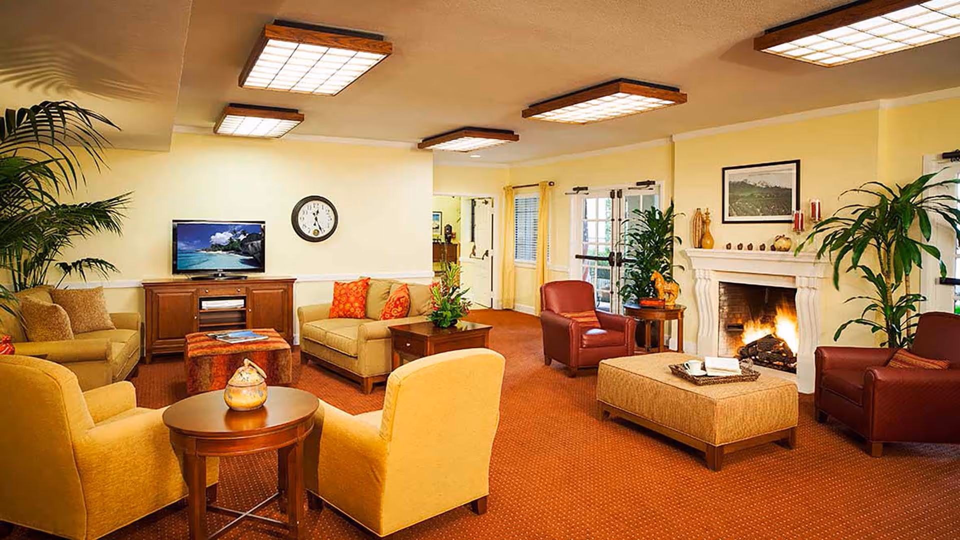 A cozy living room with beige and red armchairs, a beige sofa with red cushions, a wooden coffee table, a fireplace with a fire burning, potted plants, a wall clock, and a TV on a wooden cabinet. The room has yellow walls and a carpeted floor.
