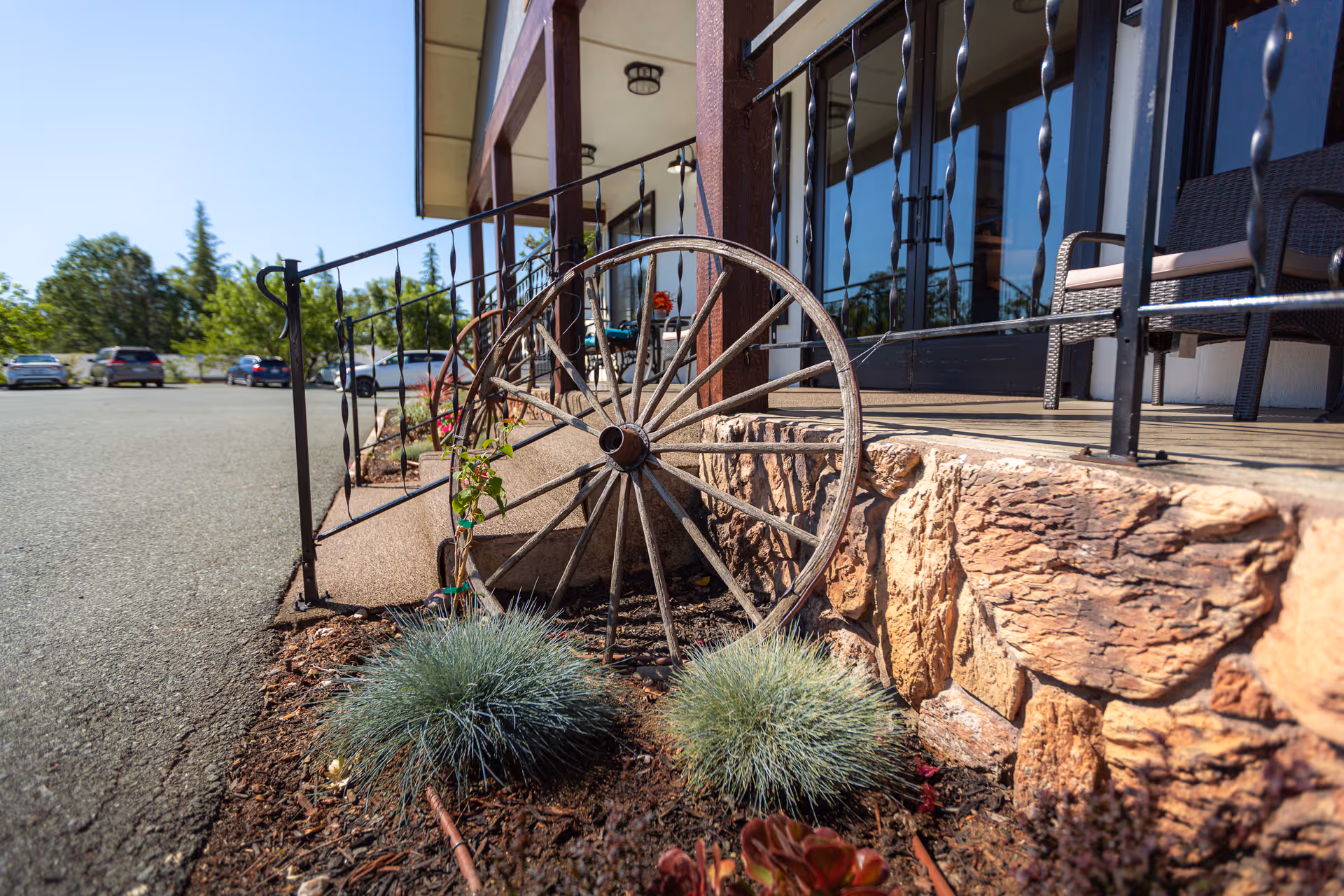 A stone-faced front porch area with a decorative wooden wagon wheel, small ornamental plants, metal railing, and a parking lot beyond.