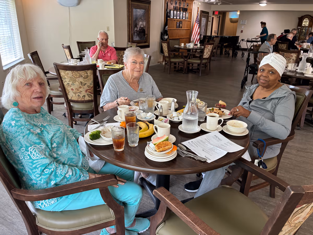 Three elderly women sitting around a round dining table in a senior living facility, enjoying a meal with plates of food, drinks, and a menu on the table. The room has multiple tables and chairs, with other residents and staff visible in the background.