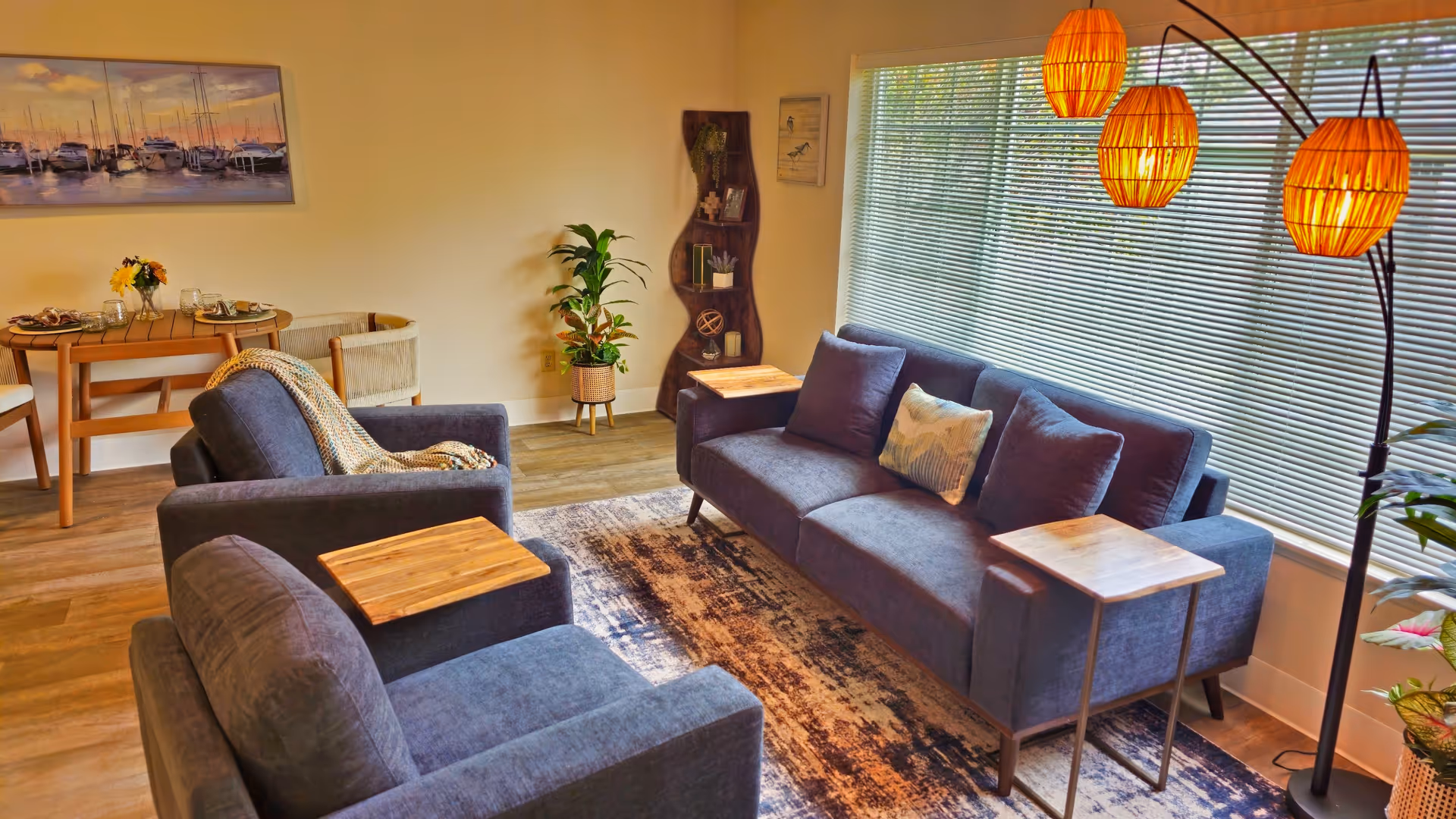 Bright furnished living room with blue sofas and armchairs, wooden side tables, a patterned rug, floor lamp, and dining table by a large window.