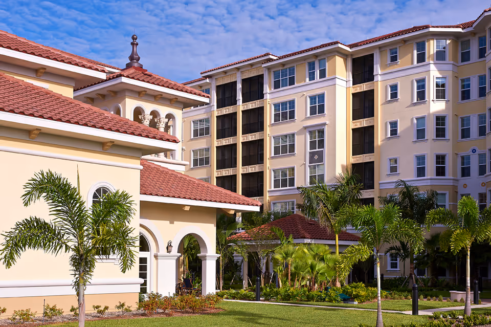 Exterior view of a senior living facility with beige buildings featuring red tile roofs, multiple windows, and arched doorways. The landscaped grounds include palm trees, green grass, and shrubs under a partly cloudy blue sky.