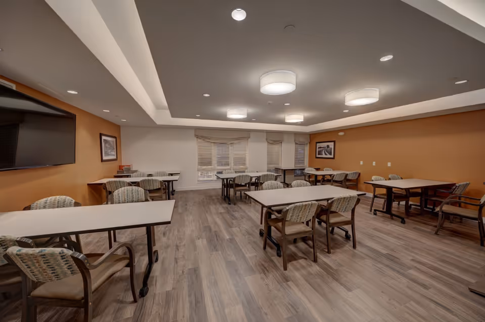 A spacious dining room with multiple tables and chairs arranged neatly. The room has wood flooring, beige and orange walls, a large flat-screen TV mounted on one wall, framed pictures, and several ceiling lights. Windows with blinds are visible on the far wall.