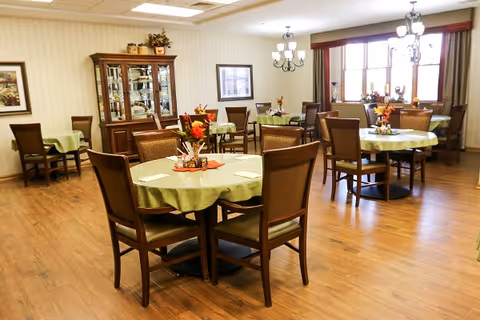A dining room with several round tables covered with green tablecloths, each set with chairs and decorated with autumn-themed centerpieces. The room has wooden flooring, a china cabinet against the wall, framed artwork, and large windows with blinds letting in natural light. Two chandeliers hang from the ceiling.