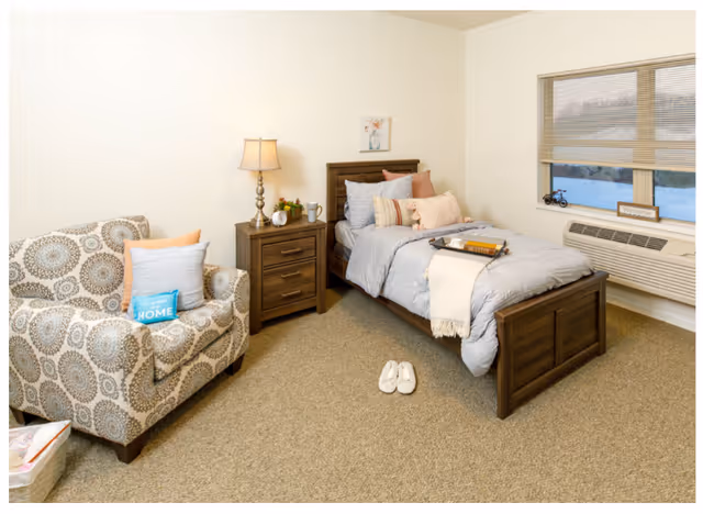 A cozy bedroom with a single wooden bed made with gray bedding and multiple pillows. A wooden nightstand with a lamp, clock, and small plant is next to the bed. A patterned armchair with decorative pillows is positioned to the left. A window with blinds and a heating/cooling unit is on the right wall. A pair of white slippers is placed on the carpeted floor near the bed.