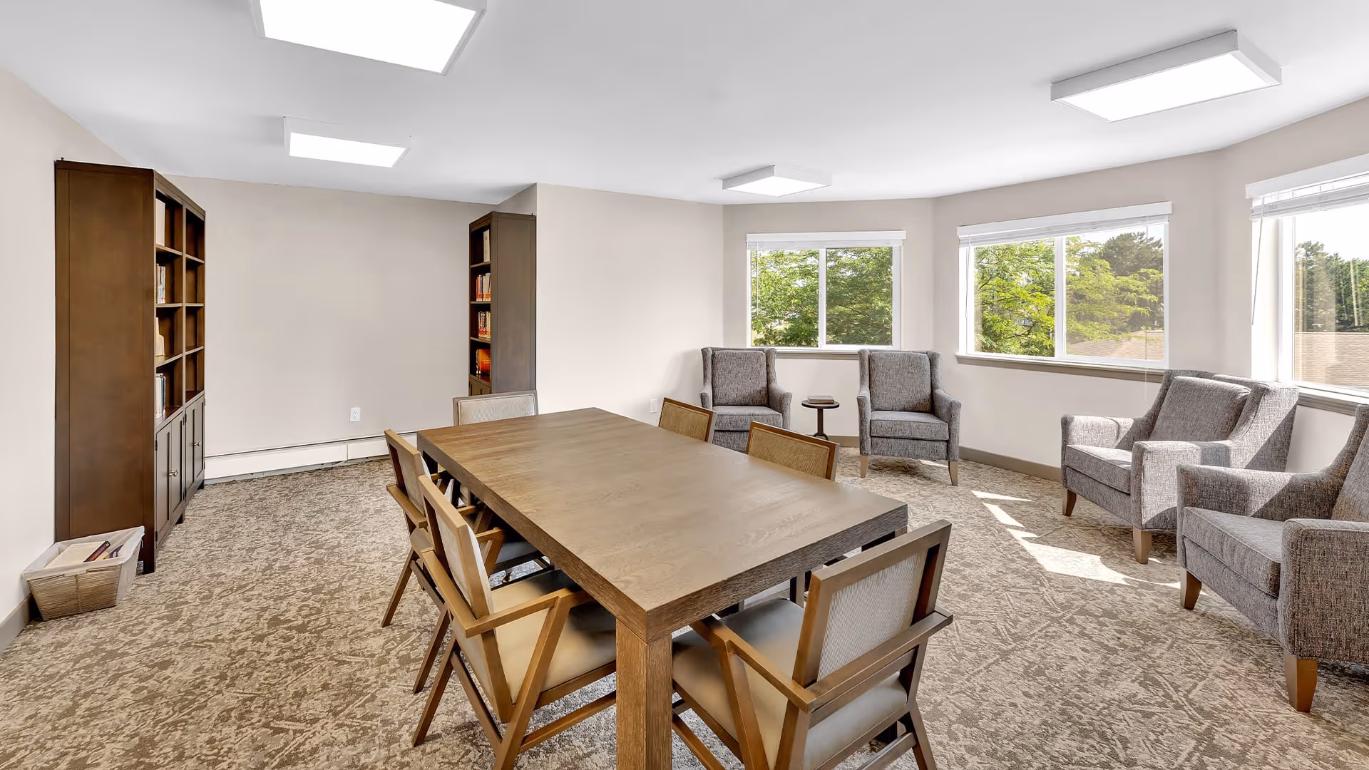 A bright room with a large wooden table surrounded by eight chairs in the center. There are four upholstered armchairs arranged near three large windows letting in natural light. Two tall wooden bookshelves filled with books are against the wall. The room has beige walls and carpeted flooring.