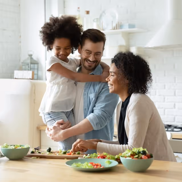 A smiling family of three in a kitchen preparing a meal together. A man is holding a young girl who is hugging him, while a woman is cutting vegetables on a wooden table with bowls of salad around.