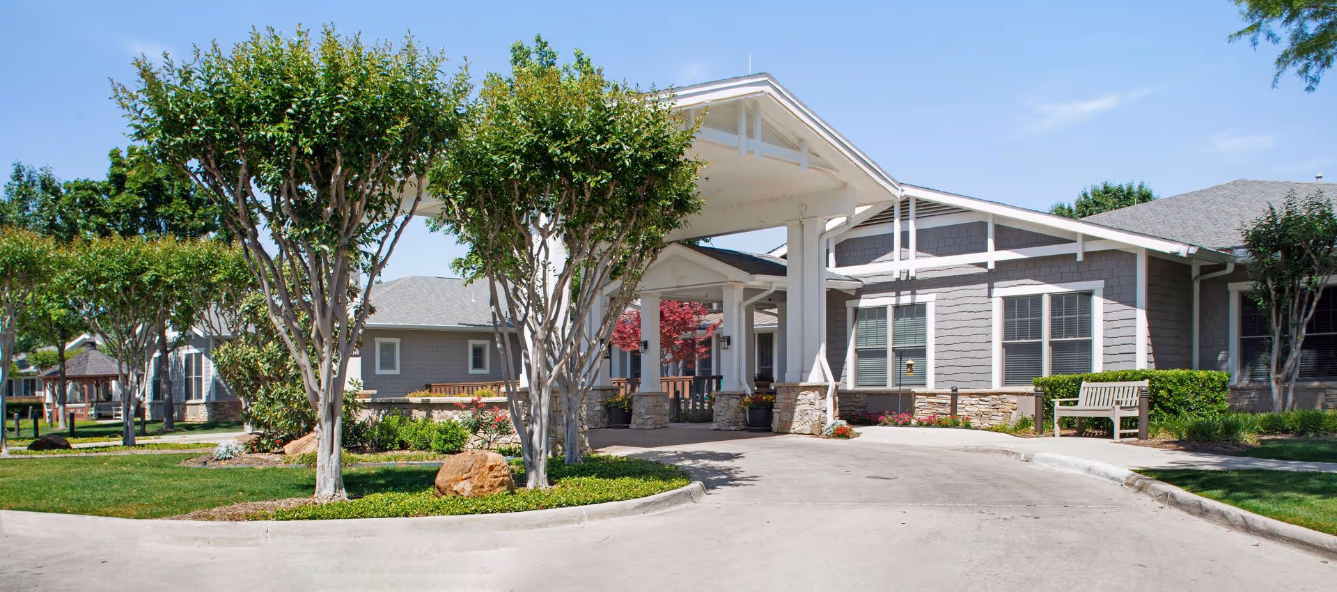 Entrance and porte-cochère of a single-story senior living facility with landscaped trees, benches, and a driveway.