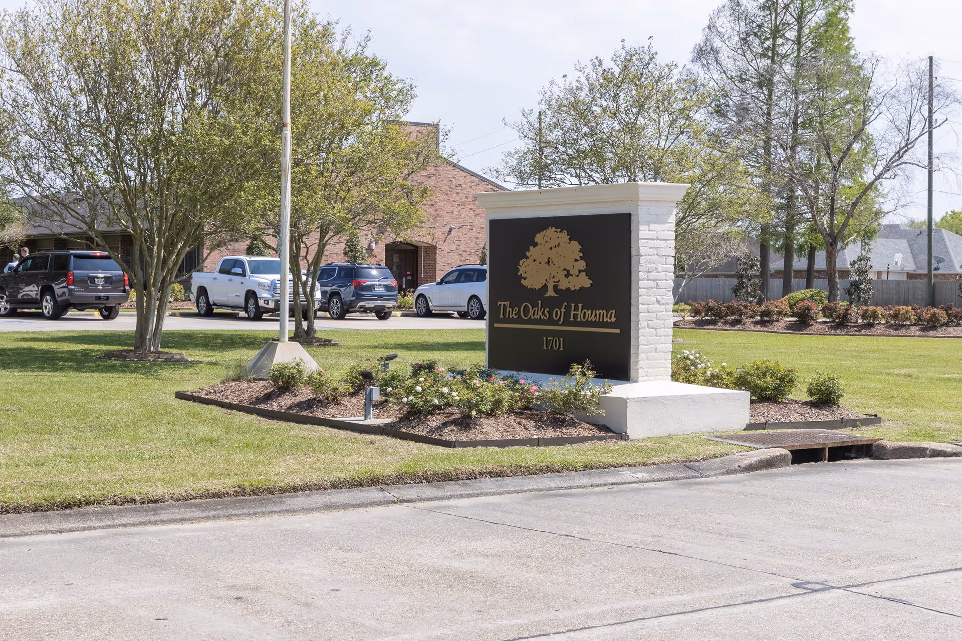 Landscape entrance sign reading "The Oaks of Houma" on a grassy median with trees, parked cars, and the facility building in the background.