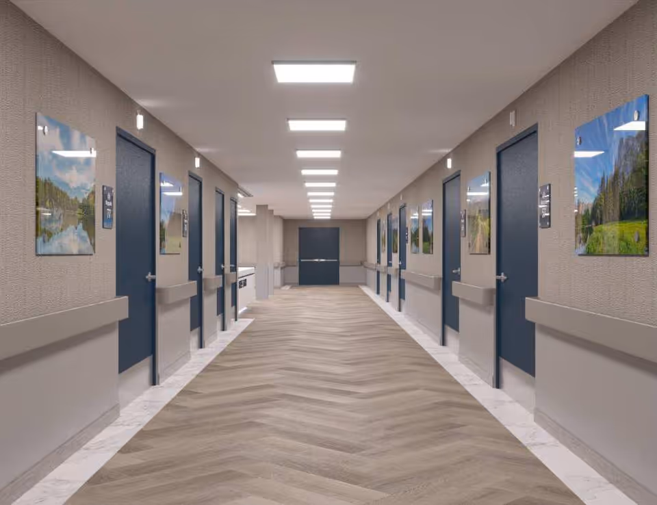 A long, well-lit hallway in a healthcare or rehabilitation facility with multiple closed blue doors on both sides. The walls are decorated with landscape photographs, and there are handrails along both sides of the hallway. The floor has a wood-patterned design, and the ceiling has evenly spaced rectangular lights.
