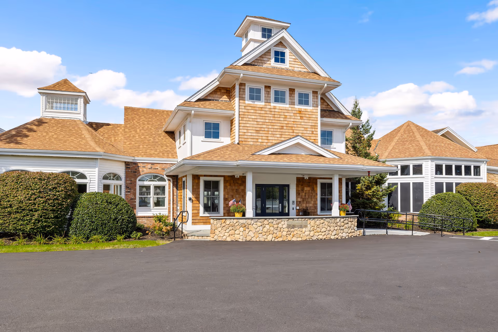 Front entrance of a multi-story senior living building with shingle siding, a stone porch, and manicured bushes under a blue sky.