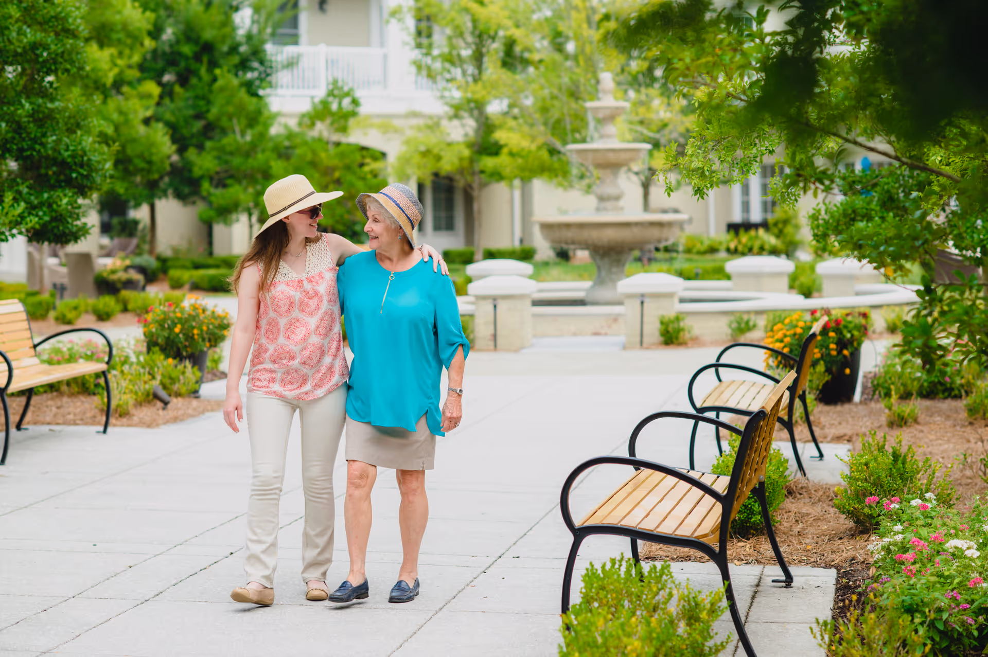 Two women, one younger and one older, walking arm in arm along a paved pathway in a garden area with benches, greenery, and a large fountain in the background. Both women are wearing sun hats and smiling at each other.