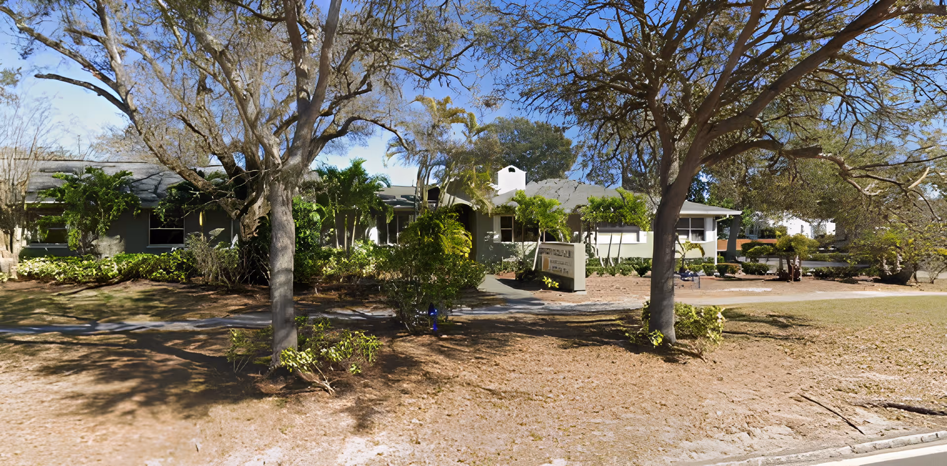 Exterior view of a single-story assisted living facility building surrounded by trees and shrubs under a clear blue sky.
