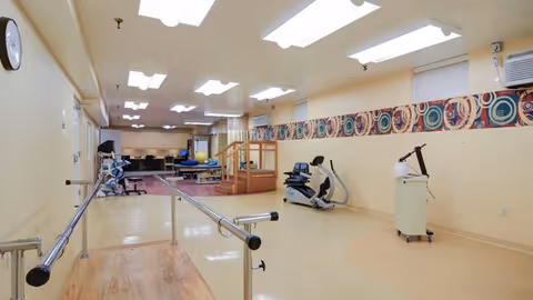 A spacious rehabilitation room with parallel bars for walking exercises, an exercise bike, therapy equipment, and a small wooden staircase with handrails. The room has bright overhead lighting, beige walls with a decorative border, and a clock on the wall.
