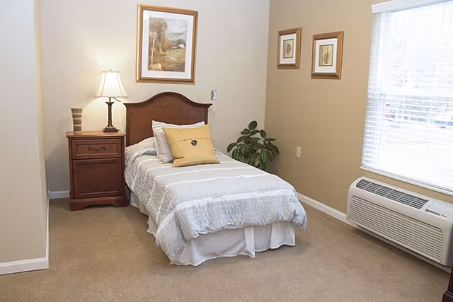 A tidy single bedroom with a twin bed, nightstand and lamp, framed artwork, a window and wall-mounted air conditioner.