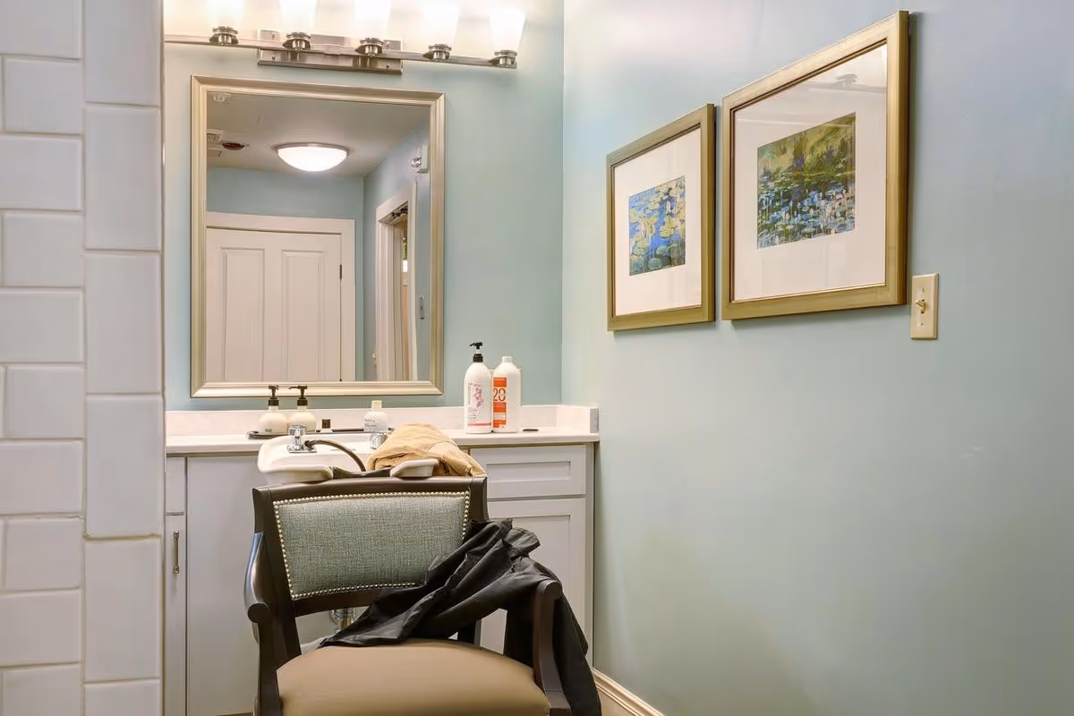 Interior view of a salon area in a senior living facility with a chair facing a sink and countertop. The wall is painted light blue and decorated with two framed paintings. A large mirror with lights above it reflects a hallway with a closed door.
