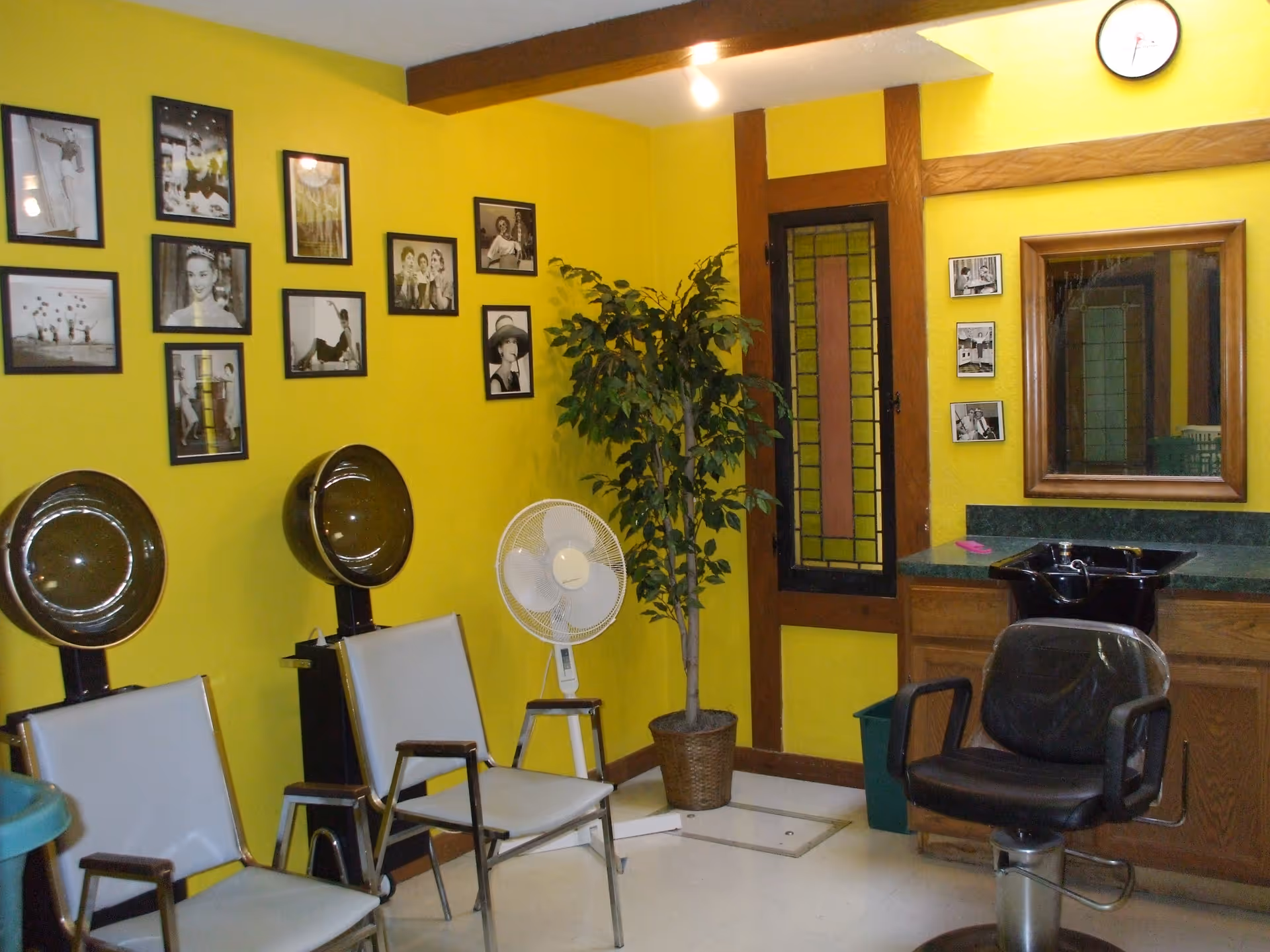 Interior of a hair salon room with bright yellow walls, two vintage hair drying chairs with hooded dryers, a black salon chair in front of a sink and mirror, a potted plant, a standing fan on a small stool, and several black and white framed photos on the walls.
