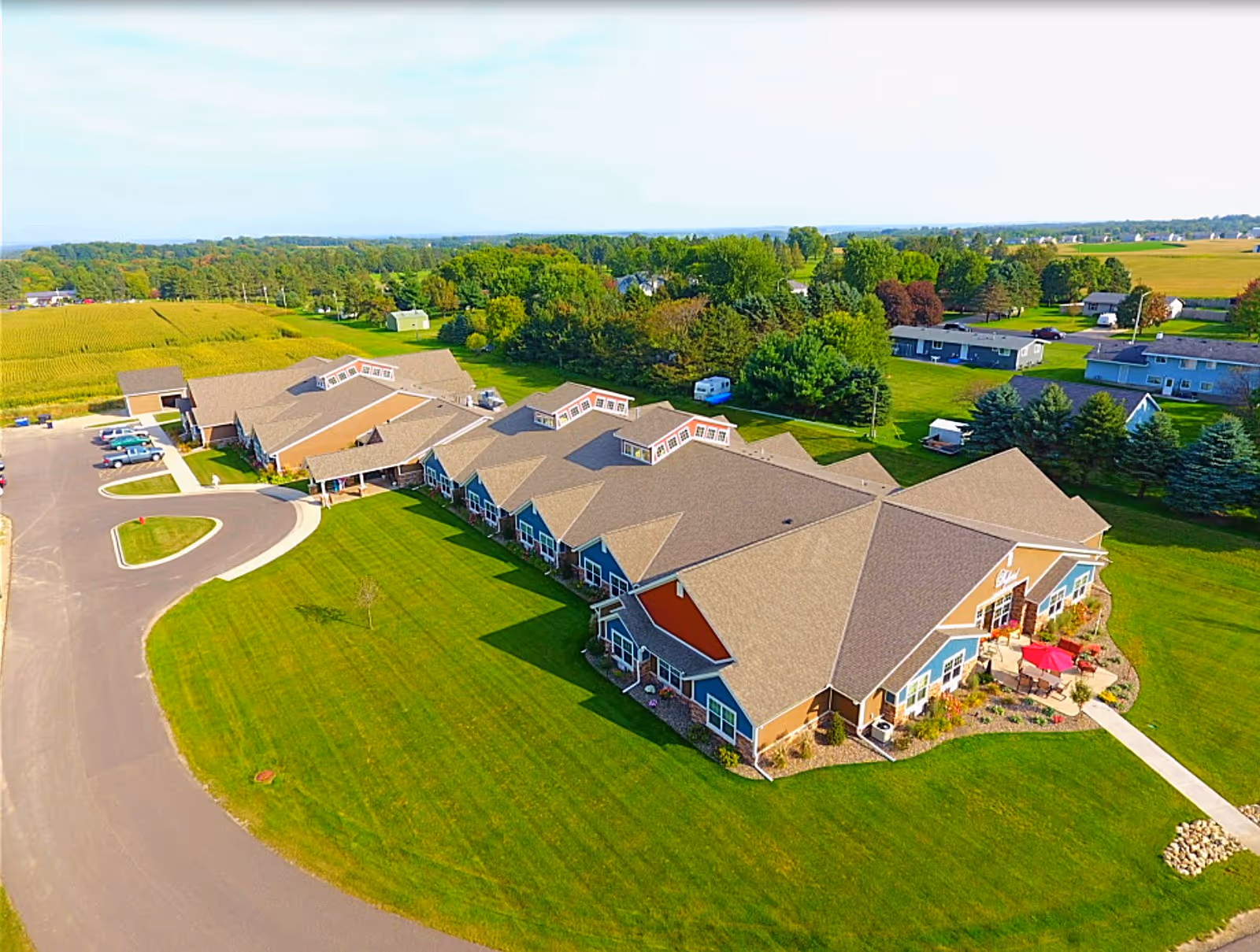 Aerial view of a large senior living facility building surrounded by well-maintained green lawns, trees, and a few parked cars. The building has a brown roof and multiple sections with windows. The surrounding area includes open fields and scattered houses.